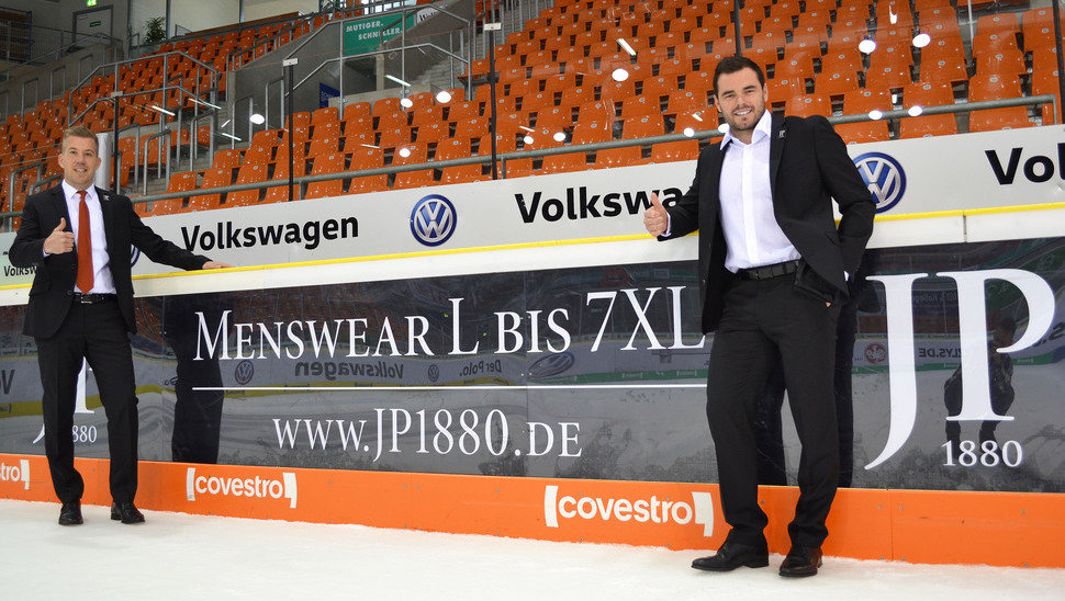 Simon Drühmel und Brent Aubin präsentieren vor der Bande von JP1880 die neuen Outfits.